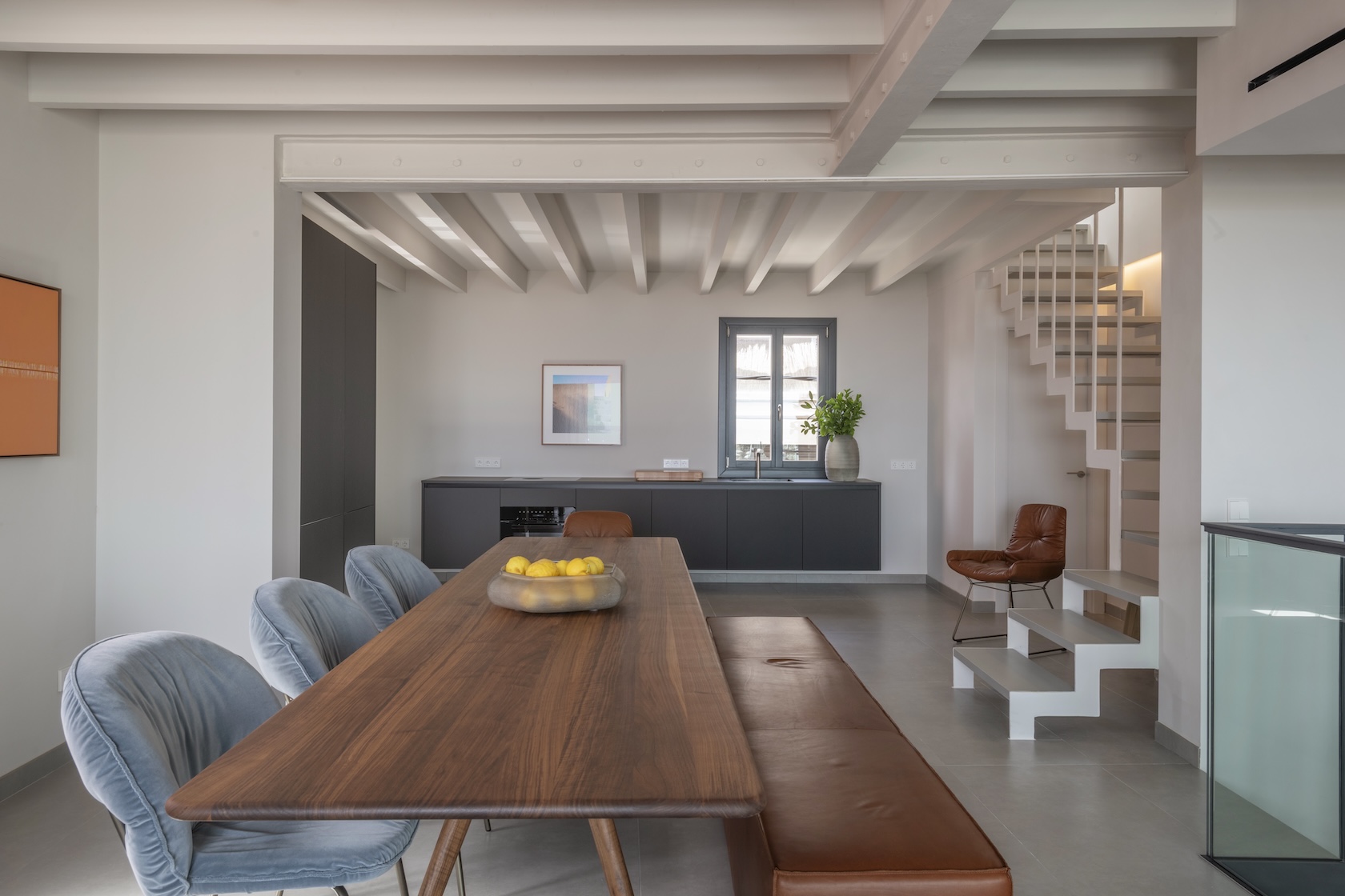 Moderner Essbereich mit Holztisch, blauen Stühlen, brauner Bank, Küchenzeile und Treppe im Hintergrund.